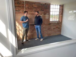 Tim Scrace and George Scott-Welsh stand inside a partitioned flexible office space with a window and exposed brick walls.